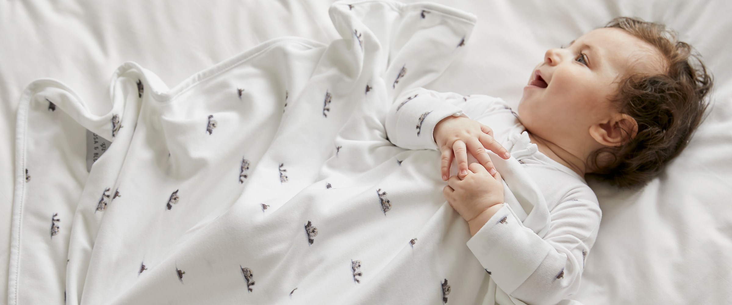 Baby in white otter printed sleepsuit laid on a white bed, with white otter printed blanket